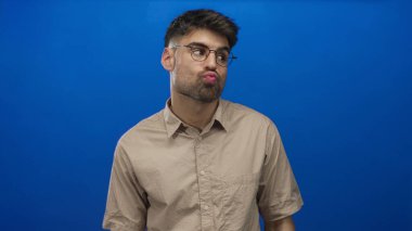 Erkekler dudaklarını büzer ve mavi duvara karşı stüdyoda yuvarlak gözlük takarak kaşlarını kaldırır..