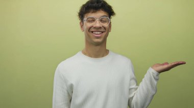 Gözlük takan ve gülümseyen beyaz süveter giyen İspanyol genç adam yeşil stüdyoda elini açık tutuyor; iyimserlik..
