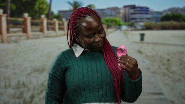 Yeşil kazaklı kadın parkta pembe kurdele tutuyor. Arka planda binalar var. Açık hava ortamında meme kanseri farkındalığı ve desteğine odaklanıyor..