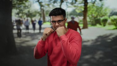 Kırmızı gömlekli bir adam, güneşli bir caddede yumruklarıyla poz veriyor. Arka planda bulanık insanlar ve ağaçlar var. Açık havada kendine güvenini ve hazırlığını gösteriyor..