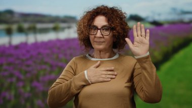 Gözlüklü İspanyol kadın, açık havada renkli bir ortamda kendine güven ve samimiyeti somutlaştıran mor çiçekler ve yeşil tarlalarla dolu canlı bir parkta yemin jesti yapıyor..