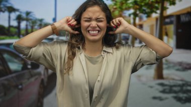 Kalabalık bir şehir sokağında kulakları çınlayan İspanyol kadın park halindeki arabalar ve ağaçlarla çevrili, açık hava ortamında rahatsız olduğunu ifade ediyor..