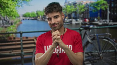 Kanal kenarındaki bisiklet parkı ve nehir kenarındaki binalarla kırmızı cankurtaran gömleği giyen adam ellerini göğsüne yaklaştırıyor..
