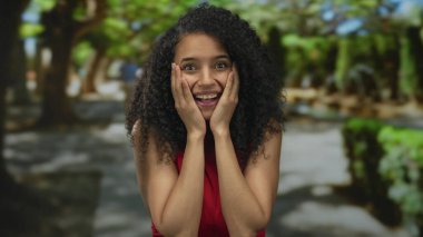 Yeşil bir parkta gülümseyen bir kadın, kırmızı bir gömlek giyiyor, kıvırcık saçları güneş ışığıyla aydınlatılıyor ve ağaçlarla çevrili, neşe ve huzur dolu bir anı yakalıyor..