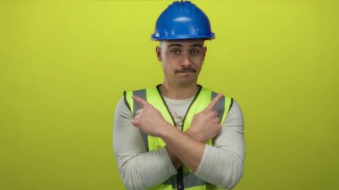 Yansıtıcı yelek giyen ve kasketi parmağıyla izole edilmiş sarı arka plan duvarını işaret eden İspanyol genç adam, güvenlik odaklı bir inşaat konseptini gösteriyor..