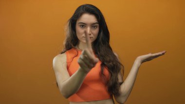 Dalgalı kahverengi saçlı, elini uzatan ve turuncu stüdyo ortamında parmağını gösteren genç bir kadın. İnkar..