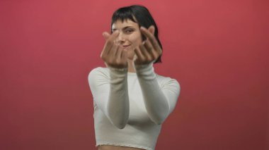 Genç İspanyol kadın kırmızı stüdyoda elleri dövmeli parmaklarıyla kalp hareketi yaparken gülümsüyor..