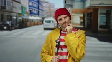 Çizgili tişörtlü ve sarı ceketli genç İspanyol bir adam elinde olta tutan bir şehir caddesinde duruyor, kırmızı bir şapka takıyor, açık havada düşünceli bir görünüm sergiliyor..