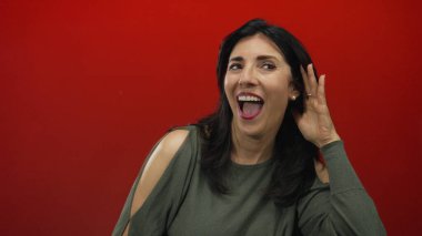 Sağ kulağını kavrayan ve stüdyoda öne doğru eğilen parlak kırmızı arka planlı bir kadın açık bir şekilde belli belirsiz bir ses duymak için; merak.