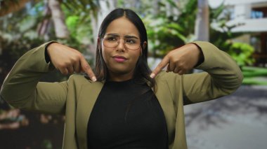 Arka planda yeşil yapraklı, gözlüklü ve zeytin ceketli, düşünceli bir ifadeyle Latin kökenli bir kadın parkın dışını işaret ediyor..