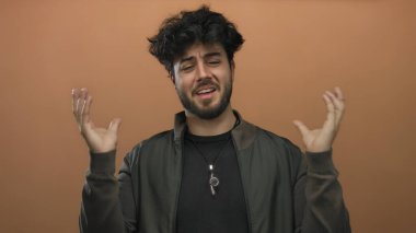 Genç sakallı İspanyol adam, portakal arka planına uzanan ellerini uzatarak kafa karışıklığı ve duygularını ifade ediyor..