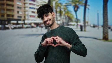 Genç adam güneşli bir şehir sokağında yeşil bir kazak giymiş, kameraya gülümseyen, sıcak ve misafirperver bir atmosfer yaratan bir kalp hareketi yapıyor..