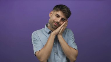 Açık mavi tişört giyen İspanyol genç adam mor stüdyo ortamında elleriyle yüzünü tutuyor..