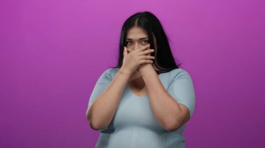 Kadın elleri pembe arka planda durarak ağzını kapatıyor, şaşkınlığını ifade ediyor, mavi gömlek giyiyor, duygularını ifade ediyor, genç, iri yarı bir kadına ait, canlı bir ortamda izole edilmiş..