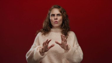 Kadın, canlı kırmızı bir arkaplan karşısında şaşırmış, rahat bir süveter giyiyor, duygularını ifade eden gözler ve ellerle yakalıyor, yaratıcı projeler için tamamen izole edilmiş..