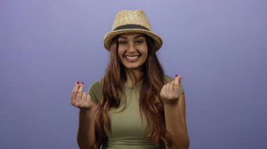 Şapkalı, gülümseyen genç bir İspanyol kadın izole bir mor arka planda parmaklarıyla para hareketi yapıyor..