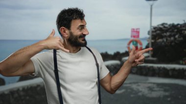 İspanyol bir adam kumsalda gezinirken jest yapıyor. Üzerinde pantolon askısı var ve gülümsüyor. Bulutlu bir gökyüzüne yaslanmış, deniz kenarında haberleşmeyi çağrıştırıyor..