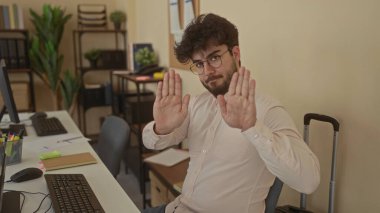 Genç İspanyol bir adam, modern mobilya ve bitkilerle dolu bir ofis odasında sakin bir ifadeyle bir masanın başında otururken duraksama hareketi yapıyor..