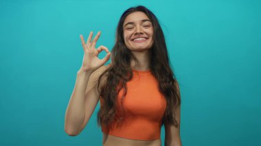 Turuncu bluz giyen bir kadın açık bir şekilde gülümserken deniz mavisi atölyesinde el işareti gösteriyor; iyimserlik..