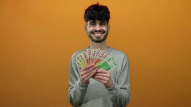 Norveç kronunu izole edilmiş turuncu arka planda tutan adam gülümsüyor, zenginlik ve mutluluk sergiliyor..