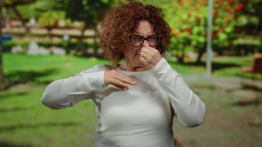 Beyaz gömlekli kadın, parkın dışında, gün boyunca yeşillikle negatif duygularını ifade ediyor. Kıvırcık saçlı ve gözlüklü hoş olmayan bir kokuyu işaret ediyor..
