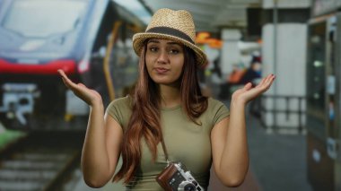 Tren istasyonunda kameralı genç bir kadın, kapalı bir yerde kapalı bir şapka takıp şehir merkezinde bulanık bir demiryolu arka planıyla şaşkın görünüyor..