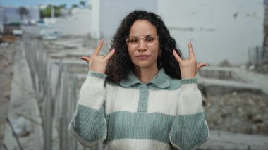 İspanyol gözlüklü kadın, şehir ortamında kendine güveni ve tarzı olan sıradan bir süveteri olan bir inşaat sahasında taş gibi bir jest yapıyor..