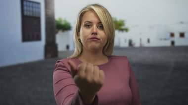 Pembe bluz giyen sarışın kadın, sessiz şehir caddesinde, beyaz bina cephesi ile avuç içiyle işaret ediyor; davet.