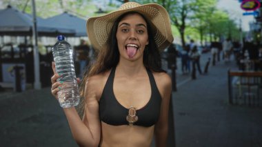 Kadın, şehir caddesinde güneş şapkası takarken dili dışarı fırlayan bir şişeyle su şişesini tutuyor; neşe.