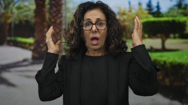 Orta yaşlı, siyahlı, şaşkın yüzlü bir İspanyol kadın arka planda palmiye ağaçları olan yemyeşil bir parkta kulaklarını kapatıyor..