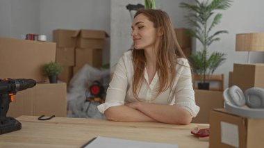 Kadın, kahverengi karton kutuların ve saksı bitkilerinin arasında ahşap masada oturuyor ve binanın içinde yumruğunu sallıyor;.