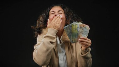 Orta yaşlı bir kadın, soyutlanmış siyah arka planına karşı Romanya banknotlarını neşeyle tutuyor, rahat süveteri ve lüleleriyle mutluluk ve şaşkınlığını dile getiriyor..