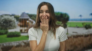 Uzun kahverengi saçlı, beyaz gömlekli, elini, yeşil bir parkta, ahşap bir köşkün yanında kapatan bir kadın..