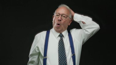 Takım elbiseli ve kravatlı kıdemli bir adam, siyah arka planda el kol hareketi yapıyor, sürpriz ve neşe ifade ediyor, gözlük, pantolon askısı ve profesyonel giysi takıyor..