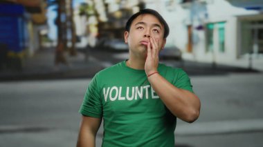 Yeşil gönüllü tişörtü giyen genç adam, arka planda bulanık binalar olan hareketli bir şehir caddesinde dikilirken düşünceli görünüyor..