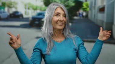 Son sınıf öğrencisi, gri saçlı, iki eli de açık bir şehir caddesinde turkuaz bir kazak giymiş ve gün ışığında hafifçe gülümseyen bir kadın..