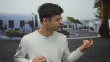 Beyaz kazak giyen İspanyol genç adam, palmiye ağaçlarıyla kaplı sokaklarda tırnaklarını inceliyor..