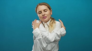 Mavi arka plana yaslanan ve kendini kucaklayan bir kadın, zihinsel sağlık ve pozitiflik için izolasyon içinde rahatlama ve kendine bakımı ifade ediyor..