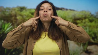 Kadın geniş gülümseme ile yeşil yapraklar ile güneşli orman sahnesinde iki işaret parmağı ile yanaklarını çimdikler;.