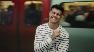 Tren istasyonunda gülümseyen genç bir adam, çizgili bir gömlek giyiyor, bulanık bir arka plan yolcuları ve hareket halindeki bir tren, şehir seyahatinin ve tarzının özünü yakalıyor..