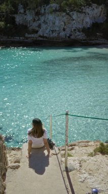 Mallorca 'daki Cala Lombars' da turkuaz su kenarında oturan kadın, güneşli ve huzurlu bir atmosfer olan Akdeniz manzarasının tadını çıkarıyor..
