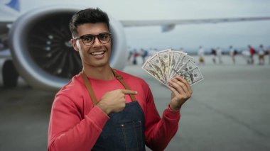 Havaalanı terminalinde bize dolar tutan ve arka planda seyahat ve zenginlik kavramını gösteren günlük giysiler içinde gülümseyen ve parayı işaret eden bir adam..