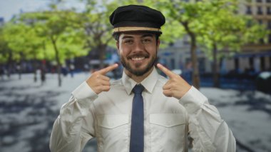 Üniformalı genç İspanyol adam, güneşli bir sokakta yeşil ağaçlar ve bulanık şehir geçmişiyle gülümseyen pozitifliğini ve sıcaklığını iletiyor..