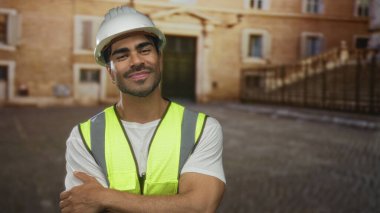 Beyaz şapka takan İspanyol bir adam ve eski bir kasaba binasının önünde kollarını kavuşturmuş sarı güvenlik yeleği giyen bir adam..