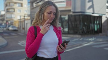 Şehir sokağında akıllı telefon kullanan bir kadın, düşünceli bir ifadeyle, pembe ceket ve beyaz gömlek giyiyor, modern şehir ortamında, genç profesyonel görünüş, çağdaş yaşam tarzı.