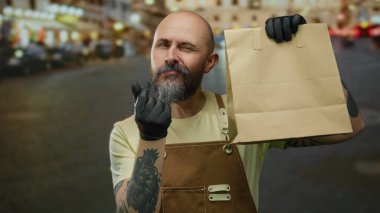 Sakallı, önlüklü ve eldivenli kel bir adam gece şehir sokaklarında kağıt torba tutuyor. Şehir ışıklarının altında benzersiz bir dövme sergiliyor..