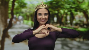 Parkta taç giyen bir kadın elleriyle kalp hareketi yapıyor, neşe ve kendini beğenmişlik gösteriyor, gür ağaçlarla çevrili, açık havada zarafet ve oyunculuğu somutlaştırıyor.