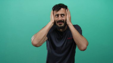 Sıkıntılı sakallı İspanyol bir adam canlı yeşil arka planda başını tutuyor, hayal kırıklığını ve kafa karışıklığını ifade ediyor..