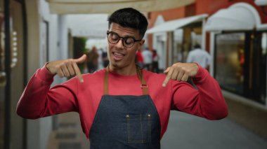 Canlı bir sokak ortamında kırmızı bir kazak ve kot önlük giymiş, kendine güveni ve modern şehir tarzı açık hava tarzıyla gülümseyen genç İspanyol bir adam..