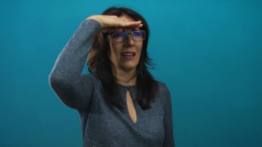 Gözlük takan bir kadın mavi stüdyoda elleri ile gözleri gölgeliyor..